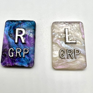 May include: Two rectangular resin tiles. One is blue, purple, and white with a silver "R" and "GRP" text. The other is white, pink, and silver with a silver "L" and "GRP" text. Both have a glittery finish.