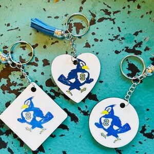May include: Keychains with blue and white bird designs. The keychains have different shapes: a square, a heart, and a circle. Each keychain has a silver chain and a tassel in yellow, blue, or black.