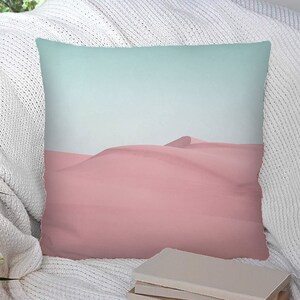 Puede incluir: Una almohada de tiro con un diseño de paisaje desértico rosa y azul. La almohada está sentada sobre una manta blanca en una silla.