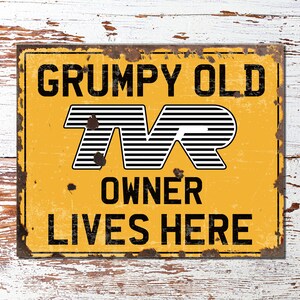 Puede incluir: Un letrero de metal amarillo con un aspecto desgastado y oxidado. El letrero dice "GRUMPY OLD TVR OWNER LIVES HERE" con un logotipo TVR en blanco y negro en el centro.