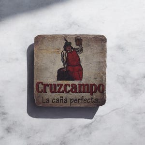 May include: A stone coaster featuring a printed image of a man in a red and white outfit holding a beer mug. The text "Cruzcampo La caña perfecta" is printed below the image.