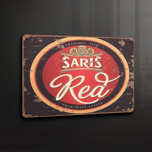 Puede incluir: Un letrero de metal de estilo vintage con un aspecto desgastado, que presenta un círculo rojo con el texto "Sariš Red" y las palabras "Prémiové Pivo" y "Polotmavý Ležiak" en una fuente dorada.