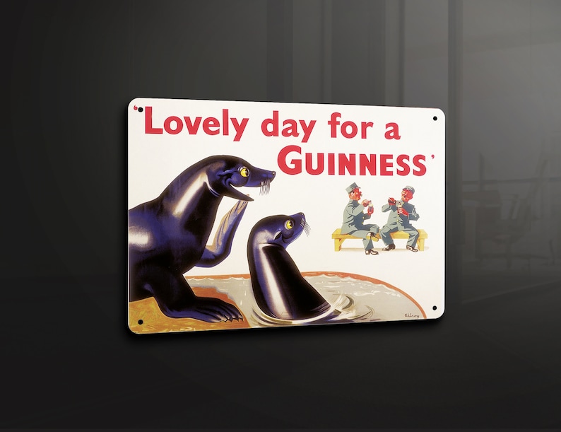 May include: A rectangular metal sign with the text "Lovely day for a Guinness" in red. The sign features illustrations of seals and two men drinking. The seals are black with yellow eyes. The men are wearing gray uniforms.