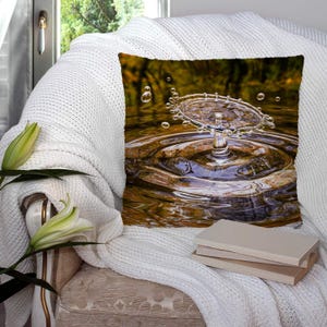 Puede incluir: Una almohada con una imagen de primer plano de una gota de agua salpicando en un charco de agua, creando ondas concéntricas. El fondo es una imagen borrosa de follaje verde.