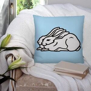 Puede incluir: Una almohada de tiro azul claro con una ilustración en blanco y negro de dos conejos acurrucados. Los conejos se miran y tienen las orejas entrelazadas.
