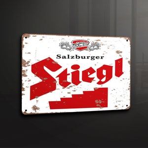 Puede incluir: Un letrero de metal blanco con un logotipo rojo y blanco para la cerveza Salzburg Stiegl. El letrero tiene un aspecto desgastado y vintage y presenta el texto "Salzburg Stiegl" en letras negras.