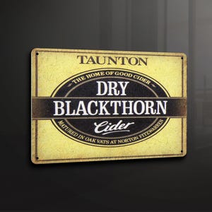 Puede incluir: Un letrero vintage amarillo y negro que anuncia Taunton Dry Blackthorn Cider. El letrero dice "The Home of Good Cider" y "Matured in Oak Vats at Norton Fitzwarren".