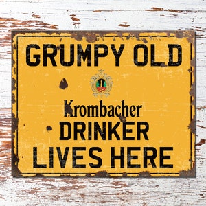 Puede incluir: Un letrero de metal amarillo con un aspecto desgastado y oxidado. El letrero dice "GRUMPY OLD KROMBACHER DRINKER LIVES HERE".