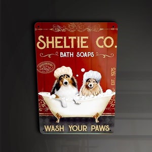 Op de afbeelding: Een rood en gouden bord met de tekst "Sheltie Co. Bath Soaps" en een afbeelding van twee Sheltie-honden in een badkuip met bubbels. Het bord heeft ook de tekst "Handmade Soap" en "Est. 1975".