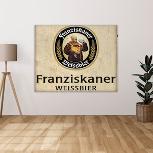 Puede incluir: Un letrero de estilo vintage con fondo marrón y borde blanco. El letrero presenta una ilustración en blanco y negro de un monje sosteniendo una jarra de cerveza. El texto "Franziskaner Weissbier" está escrito en letras negras en el letrero.