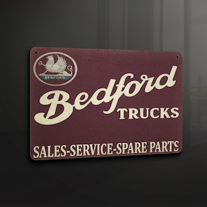 Puede incluir: Un letrero de metal de estilo vintage con fondo rojo y texto blanco. El letrero presenta un logotipo estilizado de un grifo con la palabra "Bedford" debajo. El texto "Trucks" está debajo del logotipo, y las palabras "Sales-Service-Spare Parts" están en la parte inferior del letrero.