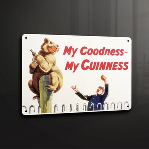 Puede incluir: Un letrero de metal blanco con un oso marrón sosteniendo una botella de Guinness y el texto "My Goodness - My Guinness". Un hombre con un uniforme azul lanza una pelota al aire.