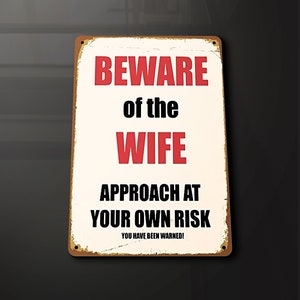 Puede incluir: Un letrero de metal blanco con bordes marrones y un aspecto desgastado. El letrero dice "Beware of the Wife Approach at Your Own Risk You Have Been Warned!"