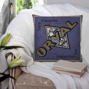 Puede incluir: Una almohada azul y dorada con un diseño vintage que presenta la palabra "Orval" en forma de diamante. El texto "trappiste" y "bier" también es visible en la almohada.