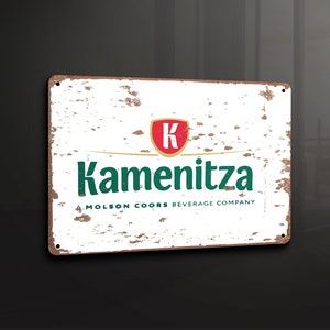Puede incluir: Un letrero de metal blanco con un aspecto desgastado y envejecido. El letrero presenta un logotipo verde y rojo con el texto "Kamenitza" y "Molson Coors Beverage Company".