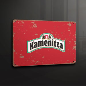 Puede incluir: Un letrero metálico rojo rectangular con la palabra "Kamenitza" en letras blancas. El letrero tiene un aspecto desgastado y envejecido con óxido visible. El logotipo incluye un borde verde y blanco con un escudo.