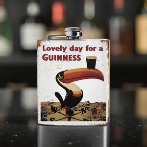 Może przedstawiać: Prostokątny metalowy flakon z zawiasową nakrętką. Flakon ma postarzane białe tło z napisem "Lovely day for a Guinness" i kreskówkowym tukanem trzymającym kufel Guinnessa. Flakon ma srebrną nakrętkę.