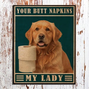 Puede incluir: Un póster de estilo vintage con un perro golden retriever sosteniendo un rollo de papel higiénico. El póster tiene el texto "Your Butt Napkins My Lady" en una fuente marrón.
