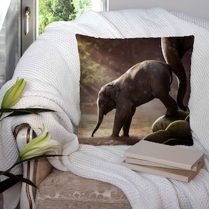 Puede incluir: Cojín cuadrado con la imagen de un elefante bebé en el agua, con la pata de otro elefante. El cojín está sobre una manta blanca de punto. El fondo incluye una ventana y flores, creando una escena tranquila.