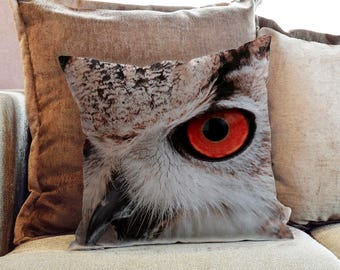 May include: A cushion with a detailed print of an owl's eye. The eye has a striking orange iris and a black pupil, with white and brown feathers. The cushion is designed to look like a close-up of the owl's face.