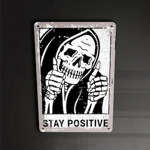 May include: A black and white illustration of a skeleton wearing a hooded robe and giving a thumbs up. The image is on a distressed, white background with the text "STAY POSITIVE."
