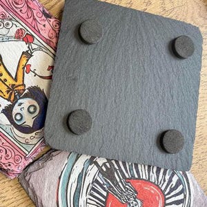 Puede incluir: Un posavasos de pizarra negra con cuatro pies de goma en la parte inferior. El posavasos es de forma cuadrada y tiene una superficie lisa.