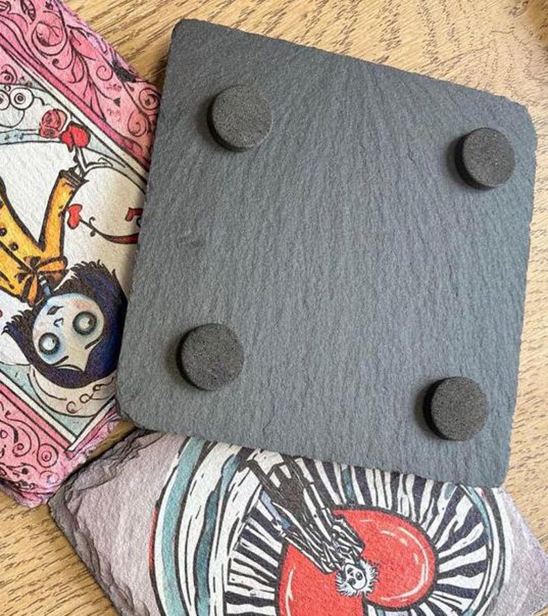 Puede incluir: Un posavasos de pizarra negra con cuatro pies de goma en la parte inferior. El posavasos es de forma cuadrada y tiene una superficie lisa.