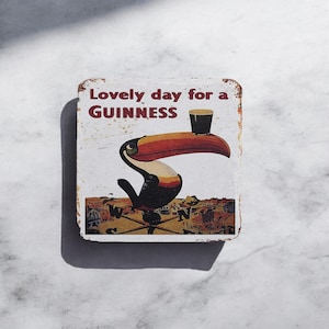 Puede incluir: Un letrero de metal blanco con un tuc&aacute;n sosteniendo un vaso de Guinness. El letrero dice "Lovely day for a GUINNESS".