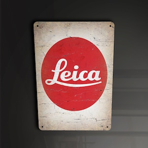 Peut inclure: Une plaque en métal de style vintage avec un cercle rouge et le mot "Leica" en lettres blanches. La plaque a un aspect vieilli et usé.