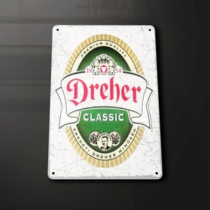 Puede incluir: Un letrero de metal blanco con un logotipo verde y dorado para la cerveza Dreher Classic. El logotipo presenta el texto "Dreher Classic" y los años "1854" y "18" en un círculo. El texto "Premium Quality" está por encima del logotipo y el texto "Eredeteti Dreher Minőség" está por debajo del logotipo.