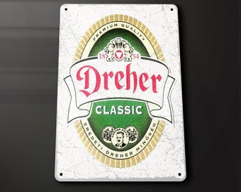 May include: A white metal sign with a green and gold logo for Dreher Classic beer. The logo features the text "Dreher Classic" and the years "1854" and "18" in a circle. The text "Premium Quality" is above the logo and the text "Eredeteti Dreher Minőség" is below the logo.