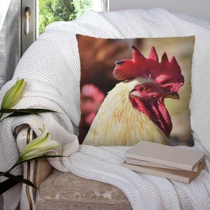 Puede incluir: Un cojín decorativo con un primer plano de la cabeza de un gallo. El gallo tiene una cresta y barbilla de color rojo brillante, con plumaje blanco crema y amarillo. El cojín está colocado sobre una manta blanca tejida sobre una silla.