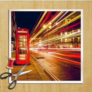 Puede incluir: Una cabina telefónica roja se encuentra en una calle de la ciudad por la noche. La calle está iluminada por las luces brillantes de los coches que pasan, creando rayas de luz en la imagen. La cabina telefónica tiene la palabra "TELEPHONE" escrita en ella.