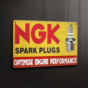 Puede incluir: Señal de metal amarilla y roja con el texto "NGK SPARK PLUGS OPTIMISE ENGINE PERFORMANCE". Una bujía blanca y plateada se muestra en el lado derecho de la señal.