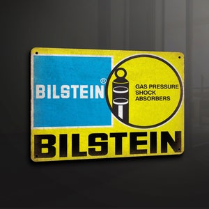 Puede incluir: Señal de metal amarilla y azul con el texto "BILSTEIN" y un gráfico de un amortiguador con el texto "GAS PRESSURE SHOCK ABSORBERS".