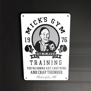 Puede incluir: Un letrero en blanco y negro con el texto "Mick's Gym 1976 Est. MCMLXXVI Training You're gonna eat lightning and crap thunder Philadelphia PA" y una imagen de dibujos animados de un hombre con bigote y un guante de boxeo.