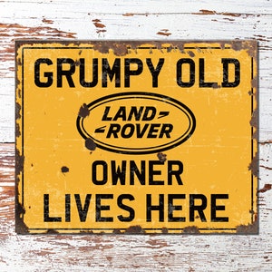 Puede incluir: Un letrero de metal amarillo con un aspecto desgastado y oxidado. El letrero dice "Grumpy Old Land Rover Owner Lives Here" con el logotipo de Land Rover en el centro.