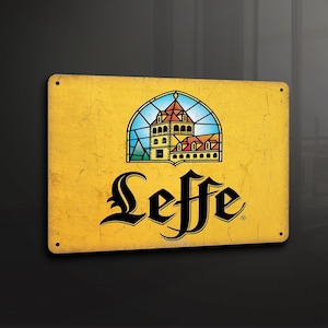 Puede incluir: Un letrero de metal amarillo con una imagen de vitral de un edificio y la palabra "Leffe" en letras negras.