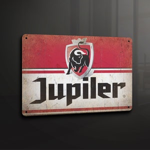 Puede incluir: Un letrero de metal de estilo vintage con fondo rojo y letras blancas. El letrero presenta un toro negro en un escudo y la palabra "Jupiler" en una fuente estilizada.