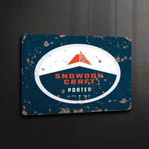 Puede incluir: Un letrero de metal de estilo vintage con un fondo azul desgastado y un círculo blanco. El círculo presenta un diseño de montaña roja y el texto "Snowdon Craft Porter 5.7% A.B.V."