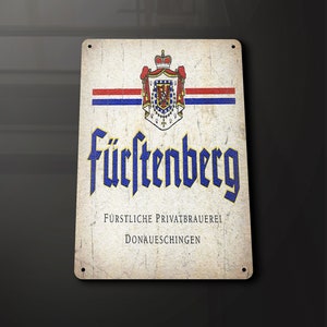 Puede incluir: Letrero metálico de estilo vintage con el logotipo de Fürstenberg. El letrero tiene un aspecto envejecido con el nombre de la marca en letras góticas azules. La parte superior presenta un escudo con una corona y rayas rojas, blancas y azules.