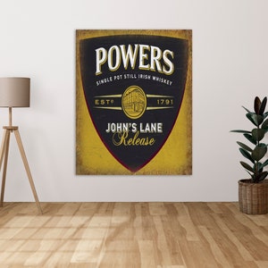 May include: A vintage-style sign with a black shield shape on a yellow background. The sign reads "Powers Single Pot Still Irish Whiskey Est. 1791 John's Lane Release".