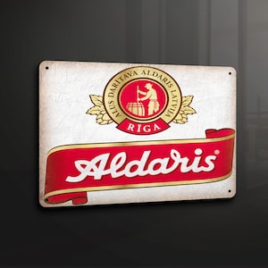 Puede incluir: Letrero metálico rectangular con el logotipo de Aldaris. El letrero presenta un fondo blanco desgastado con el logotipo de Aldaris en rojo y dorado. El logotipo incluye texto y una ilustración de una persona trabajando con un barril.