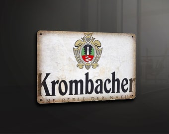 May include: A vintage-style metal sign with a faded white background and a black and white logo for Krombacher beer. The logo features a crest with a green tree and a red river, and the text "Krombacher" in a bold black font. Below the logo, the text "Eine Perle der Natur" is written in smaller black letters.