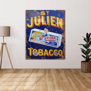 Puede incluir: Un letrero de metal vintage que anuncia St. Julien Tobacco. El letrero es azul con letras amarillas y presenta una caja de tabaco roja, blanca y azul con el texto "Ogden's St. Julien Tobacco Pure Blended Virginia".
