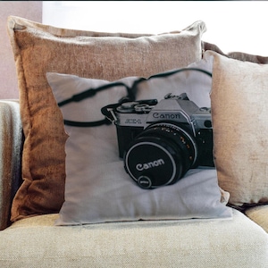 Puede incluir: Una cámara Canon AE-1 vintage de plata con una lente negra se encuentra sobre una almohada blanca y gris. La cámara tiene una correa negra y las palabras "Canon" y "AE-1" son visibles en la cámara.