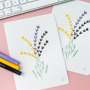 May include: Two white cards with a simple illustration of lavender flowers in purple, yellow, and black. The flowers are drawn with a simple, childlike style. The cards are on a pink surface with three markers in white, purple, and yellow.