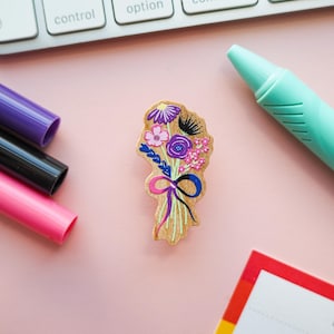 May include: A wooden bouquet charm with a pink, purple, and blue floral design. The charm is on a pink surface with a keyboard, pens, and a notebook in the background.