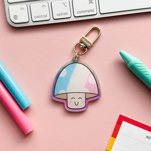 Puede incluir: Un llavero de champi&ntilde;&oacute;n rosa y azul con una cara sonriente. El llavero est&aacute; hecho de acr&iacute;lico transparente y tiene un anillo de metal dorado.
