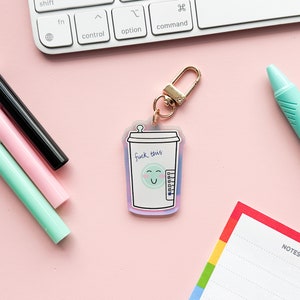 Peut inclure: Un porte-clés en or avec un charme en acrylique transparent représentant une tasse de café de dessin animé avec un visage souriant et le texte "F*ck this".